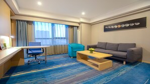 A hotel room with a desk, a blue armchair, a grey sofa, a coffee table, and a large window with curtains.