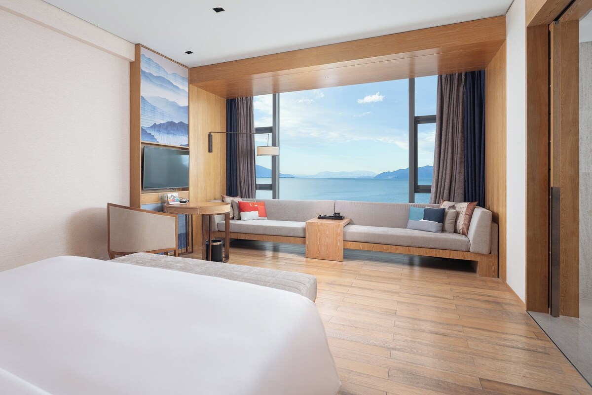 Ein Hotelzimmer mit einem großen Bett, einer Couch, einem Couchtisch und einem Fernseher. Das Zimmer hat ein großes Fenster mit Blick auf das Meer.