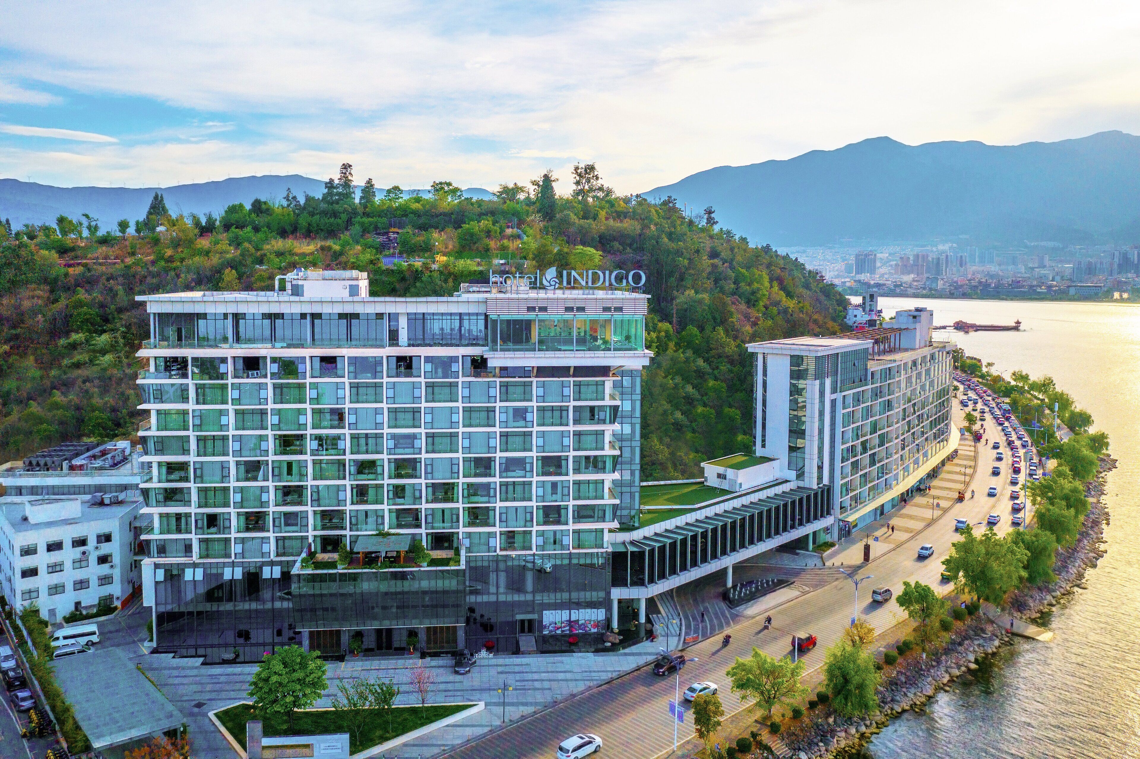 A modern hotel with multiple floors, a glass facade, and a view of a river and mountains in the background.