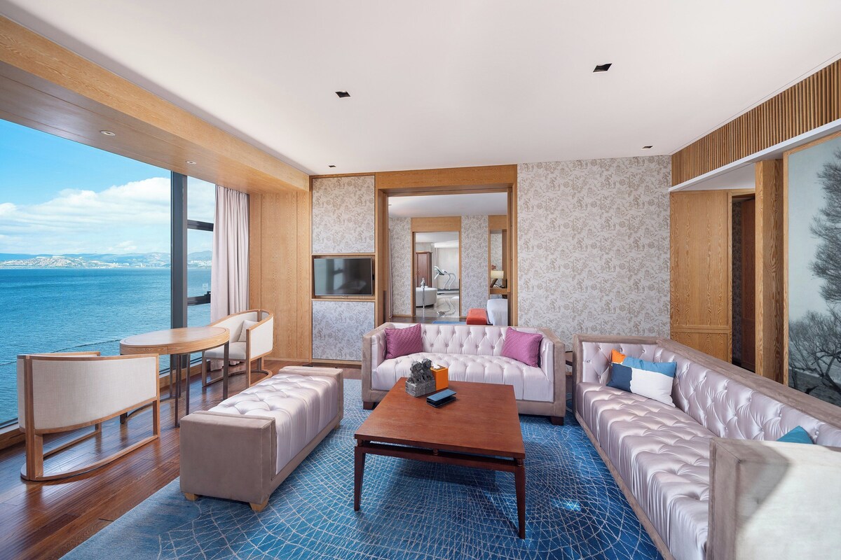 Ein modernes Hotelzimmer mit einem großen Fenster und Meerblick. Es beinhaltet ein Sofa, einen Couchtisch und eine Sitzecke mit Blick aufs Meer.