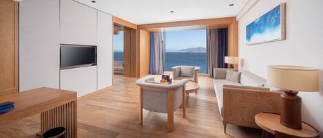 A modern hotel room with a wooden floor, a flat-screen TV, a sofa, a small table, and a view of the sea.
