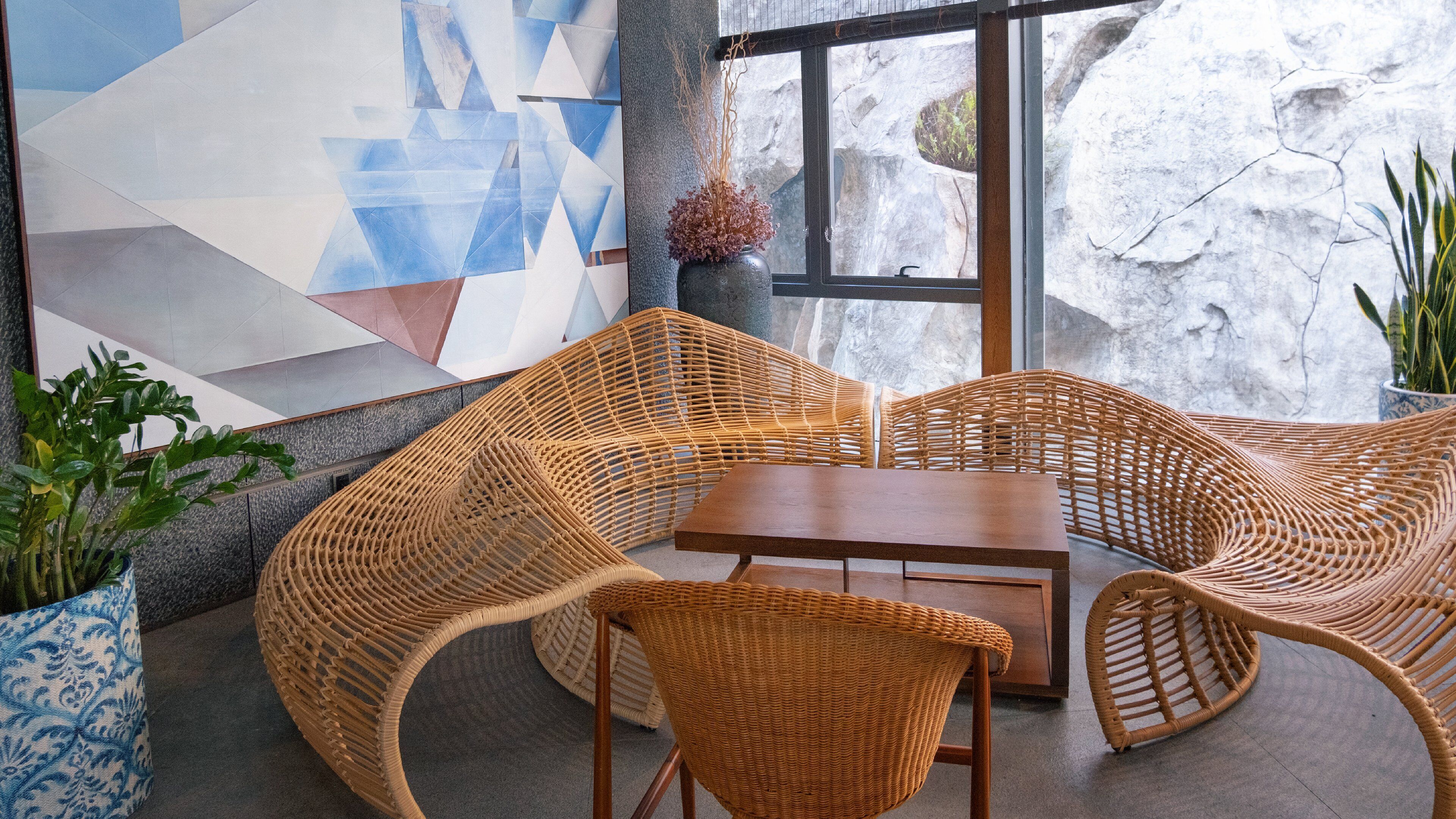 A wicker lounge area with a table and chairs, a vase with dried plants, and a geometric wall art piece.