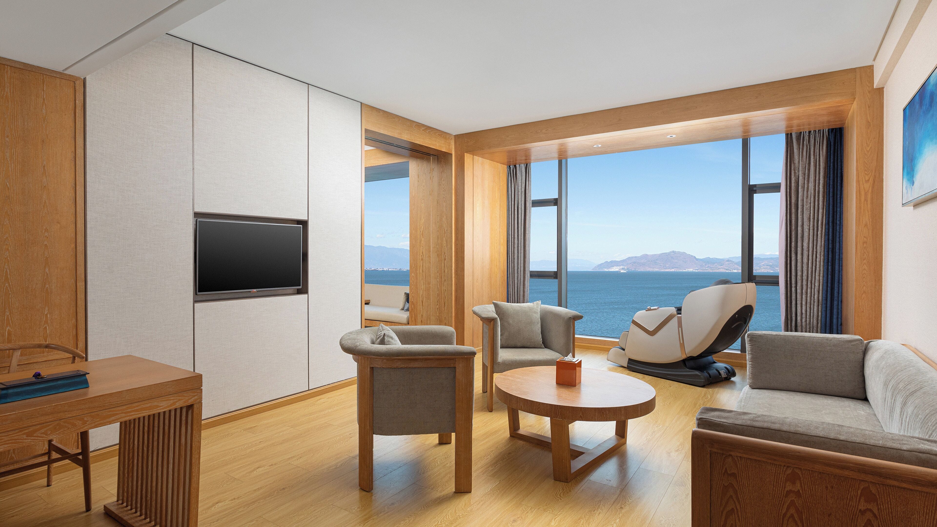 A modern hotel room with a large window offering a sea view, a flat-screen TV mounted on the wall, and a wooden desk with a chair.