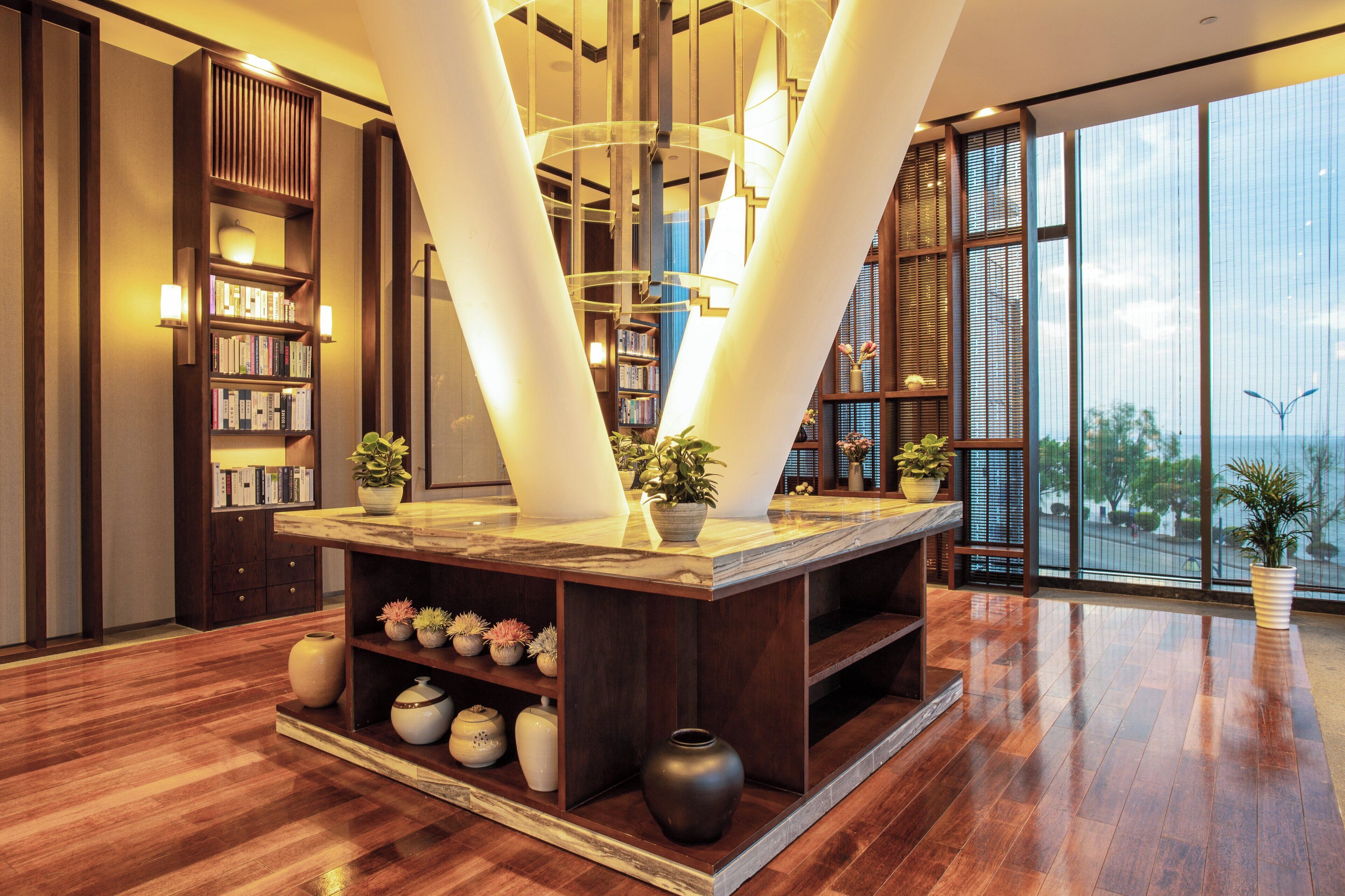 A modern lobby with a central marble table, bookshelves, and potted plants.