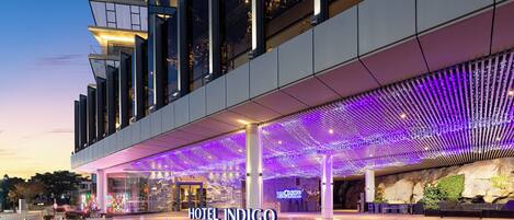A modern hotel with a prominent entrance, a covered outdoor area with a patterned ceiling, and a visible sign for Hotel Indigo.