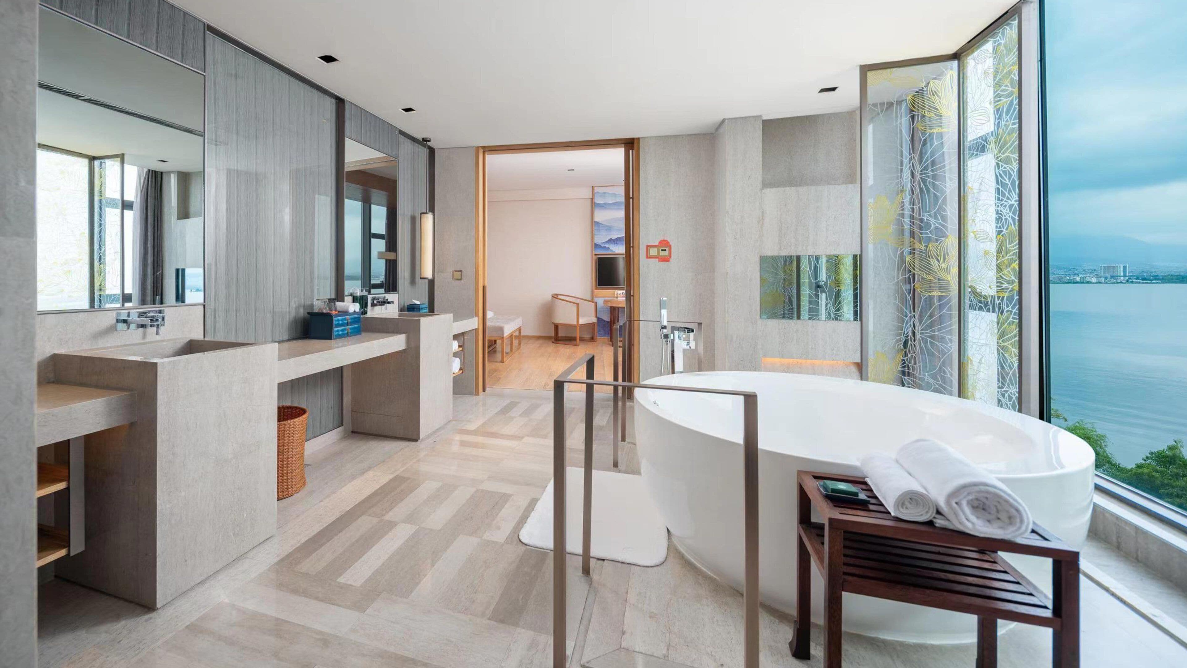 Une salle de bain moderne avec une grande baignoire, deux lavabos et un grand miroir. Les fenêtres offrent une vue sur une étendue d’eau.