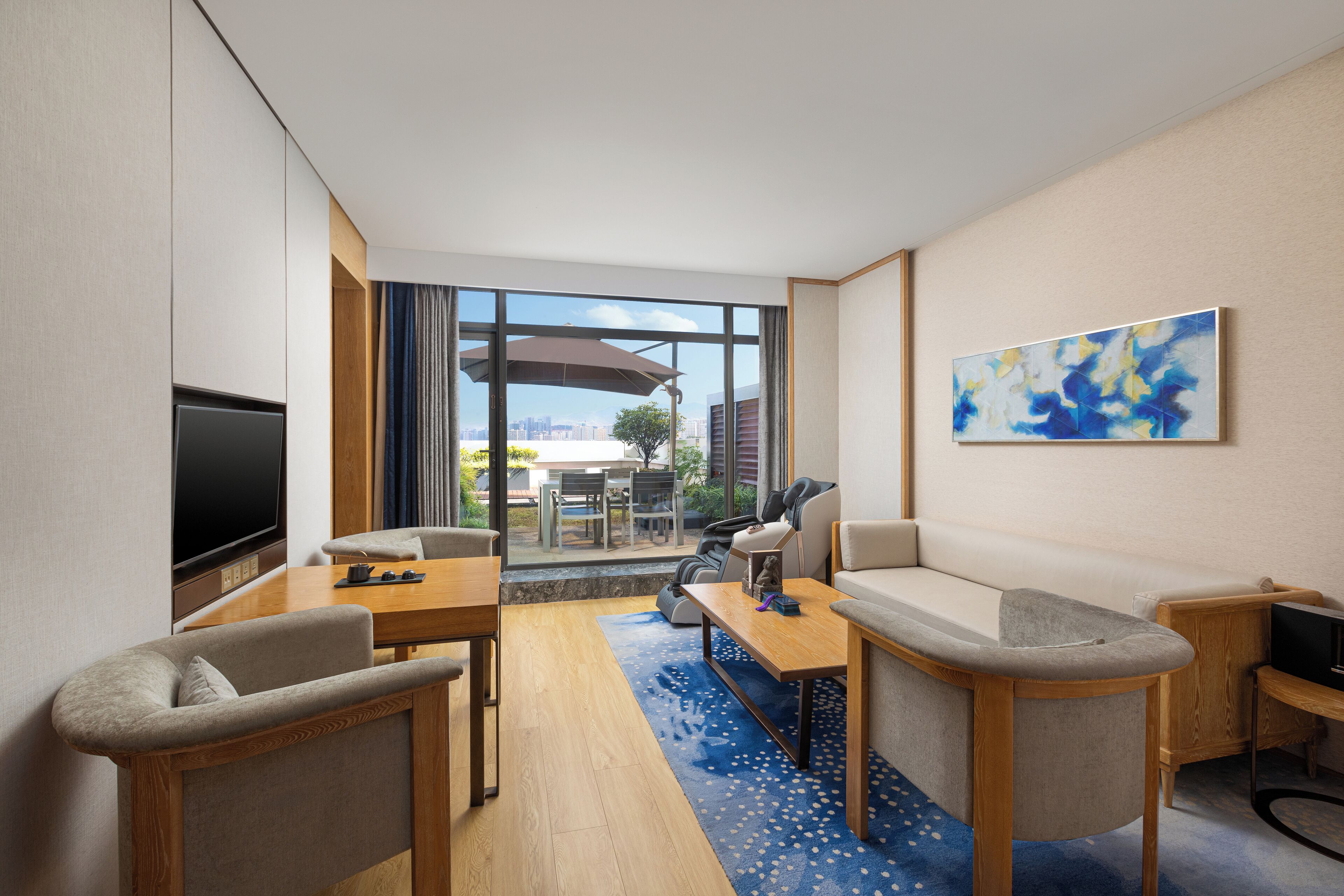 A modern hotel room with a flat-screen TV, a sofa, a small table, and a large window offering a city view.