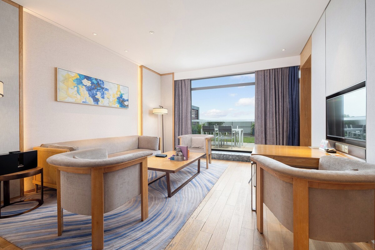 Ein modernes Hotelzimmer mit einer Couch, einem kleinen Tisch, einem Fernseher und einem großen Fenster mit Vorhängen.
