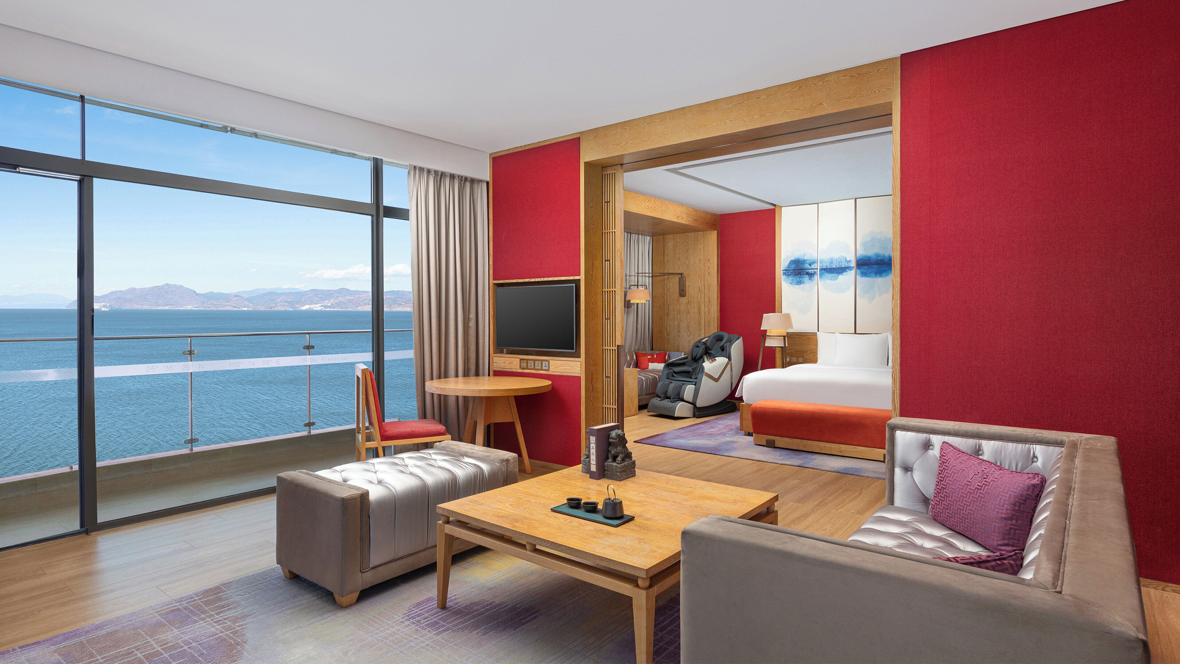 A hotel room with a large window offering a sea view, a bed, a sofa, a table, and a TV.