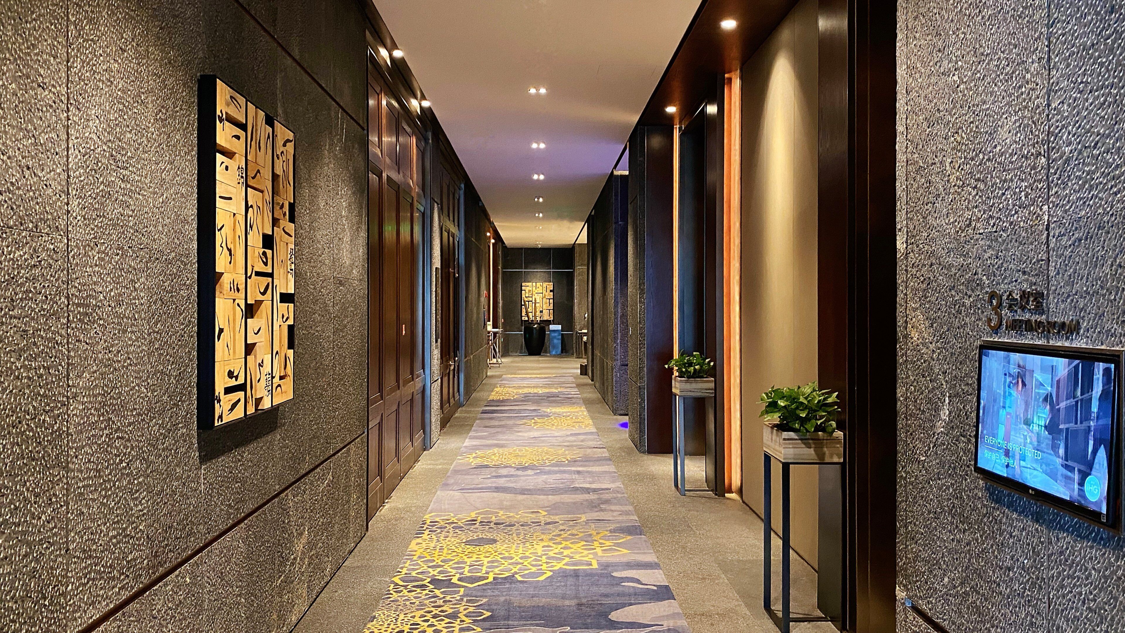 Un pasillo moderno de hotel con alfombra estampada, paneles decorativos de madera y una pantalla de televisión en la pared.
