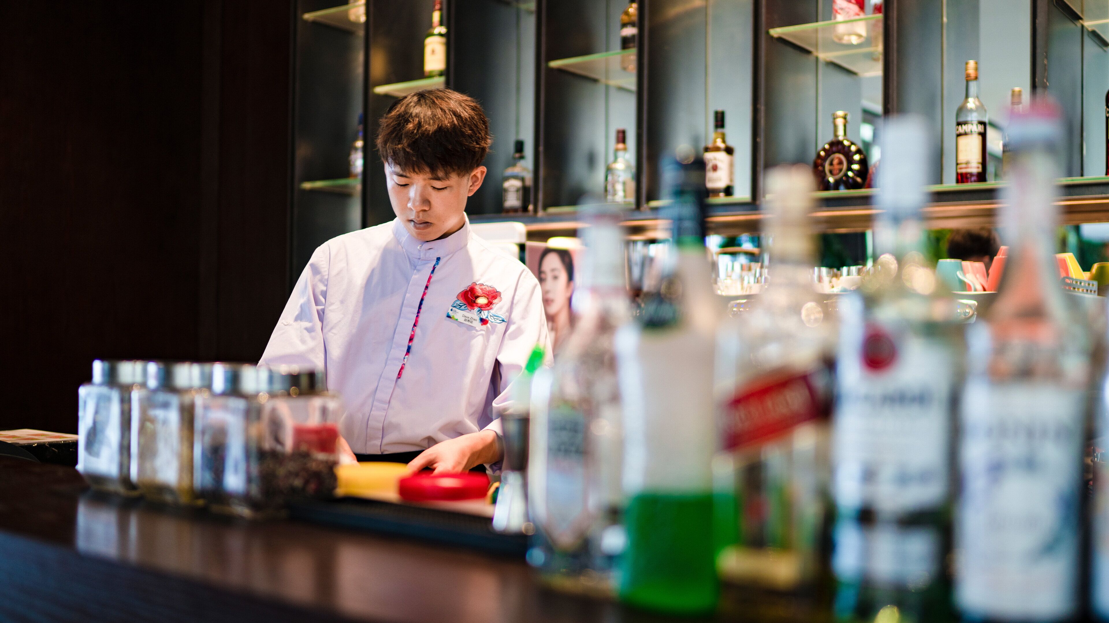 A bartender preparing drinks at a bar with shelves of bottles in the background.