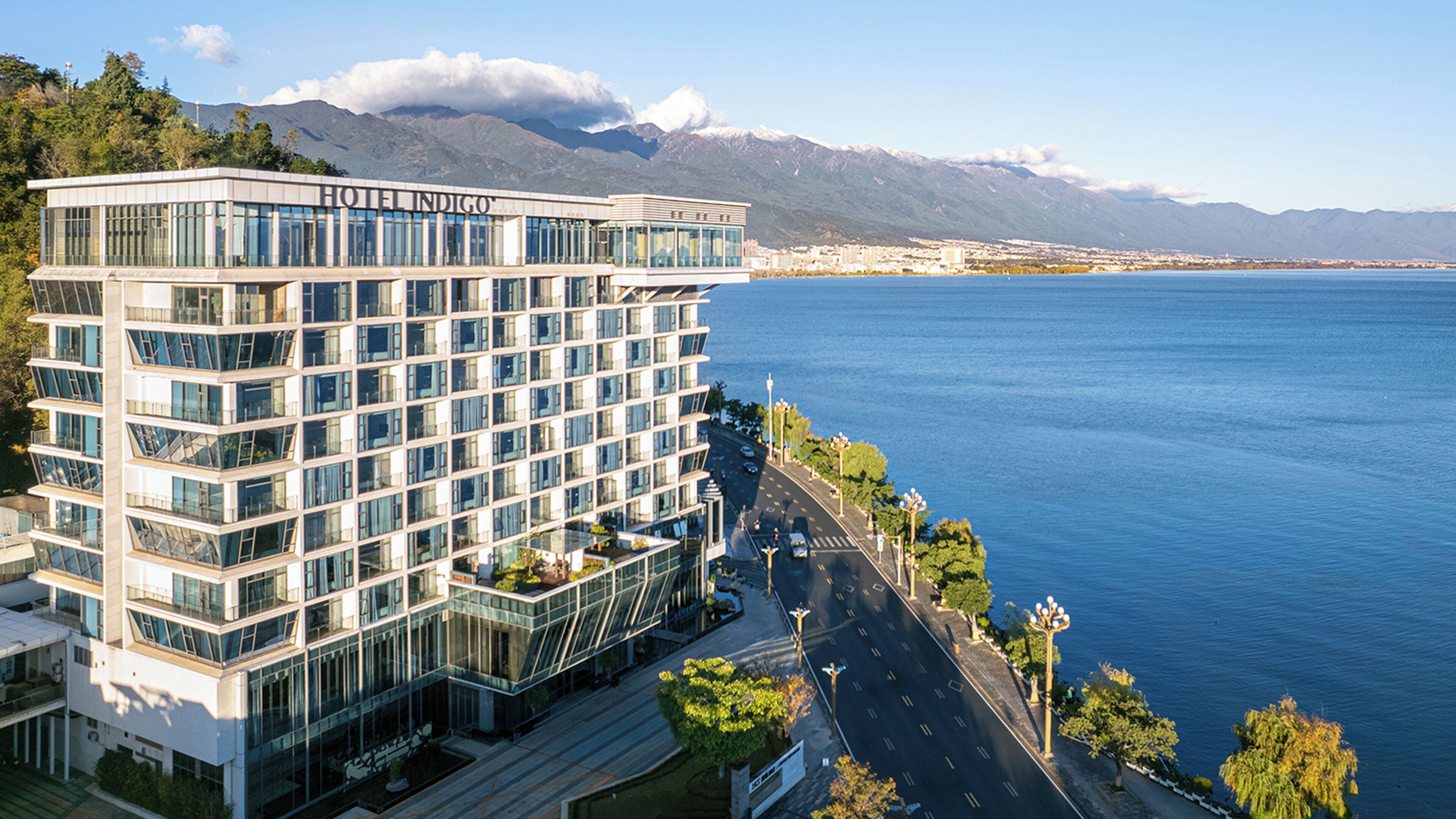 Un hotel moderno con fachada de vidrio, situado frente a la costa con montañas al fondo.