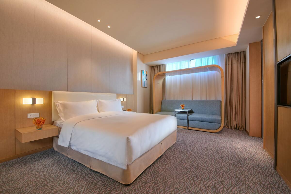 Ein Hotelzimmer mit einem großen Bett, einem kleinen Sofa, einem Nachttisch mit einer Vase und einem Fernseher.