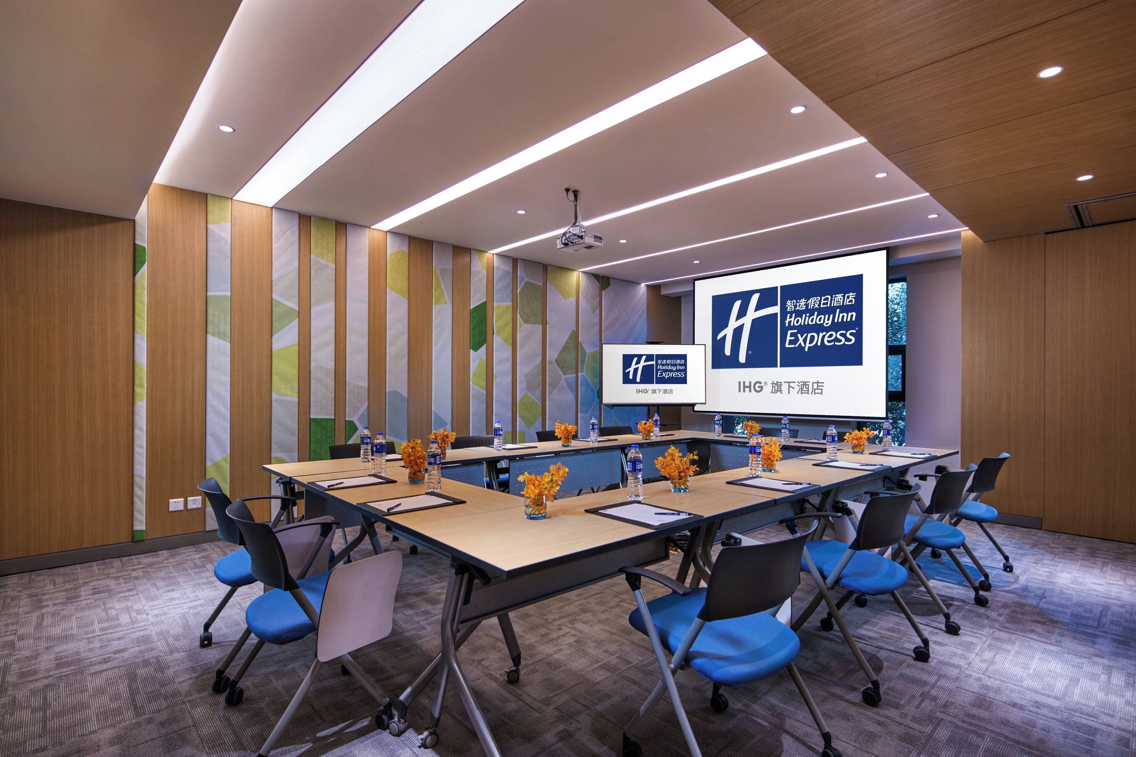 A conference room with a long table, chairs, a projector screen, and a presentation about Holiday Inn Express.