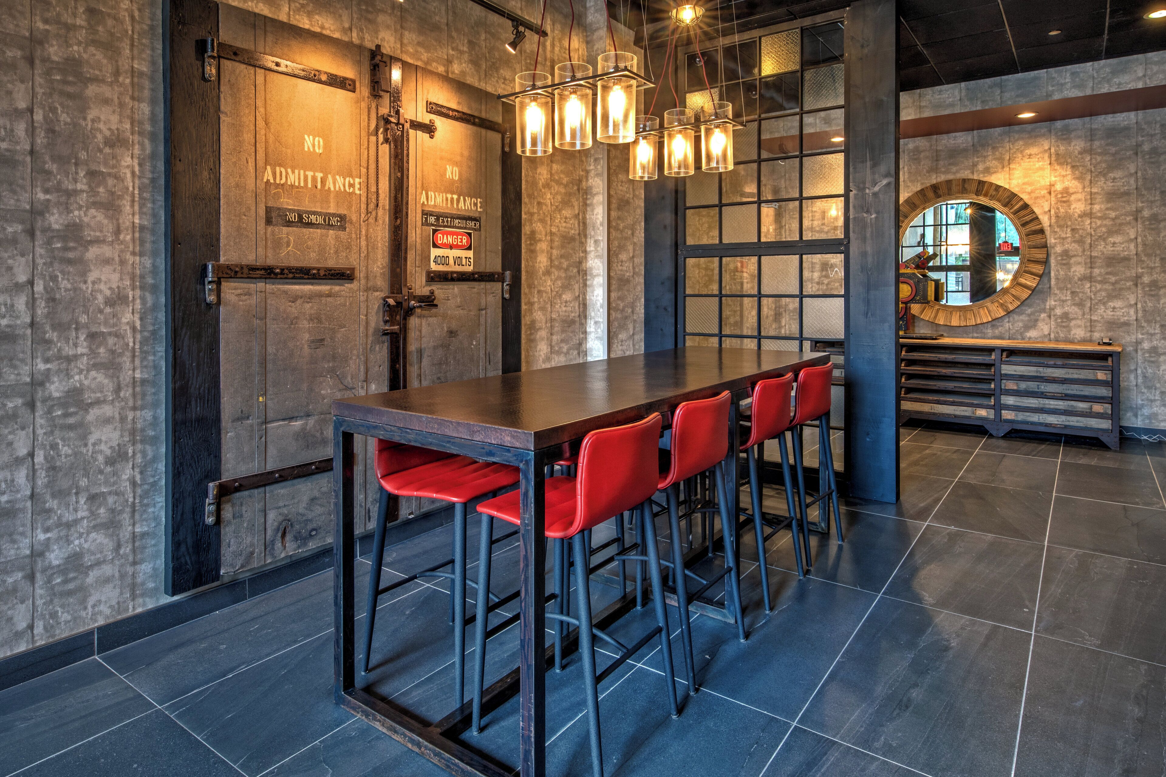 A bar area with a long table and red chairs, a wooden door with safety signs, and a round mirror on the wall.