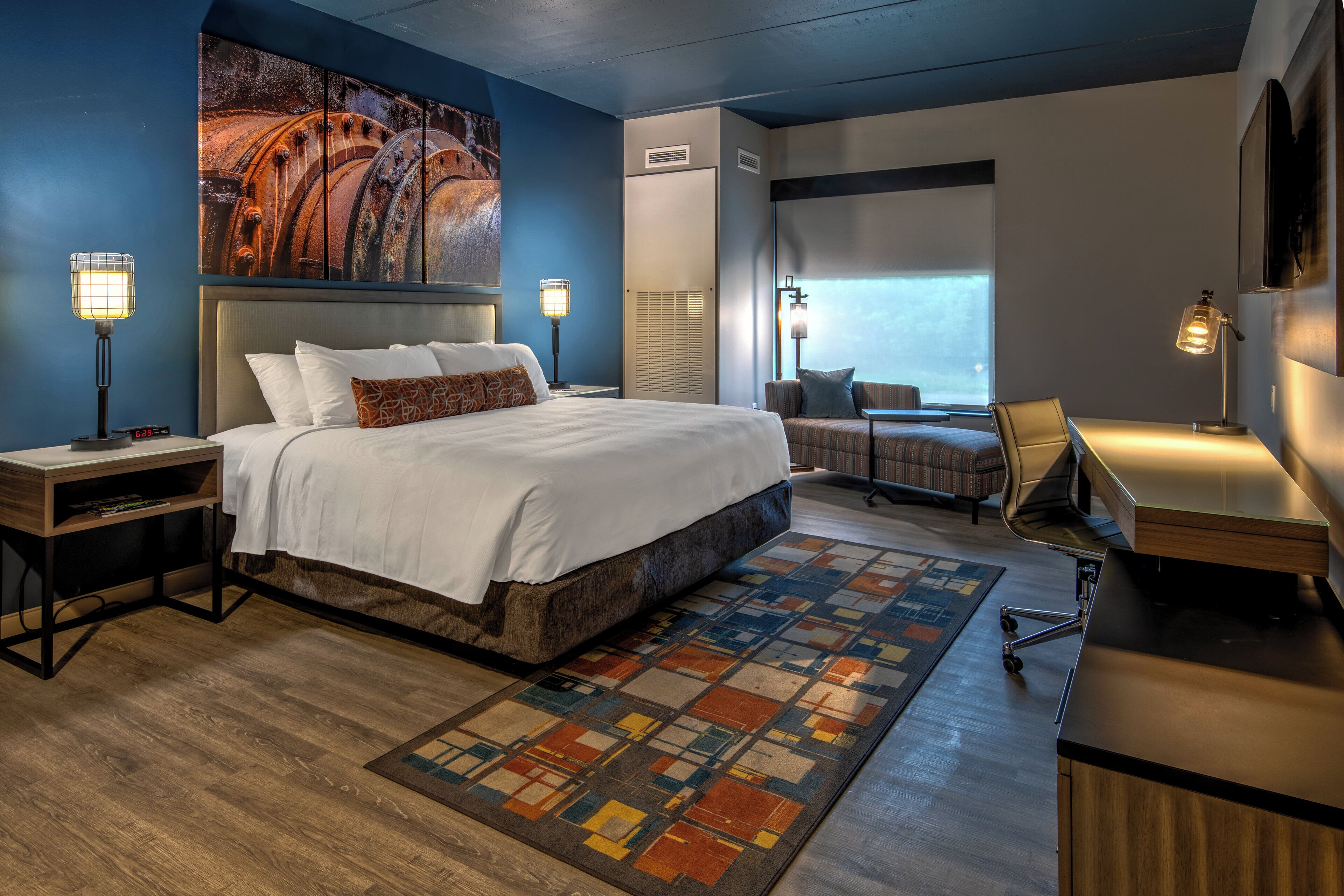 A modern hotel room with a large bed, a desk, a chair, and a view of the outdoors.