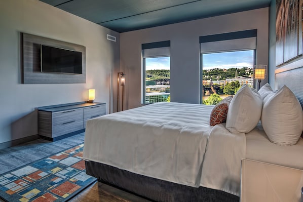 Une chambre moderne avec un grand lit, un téléviseur à écran plat et une vue sur le paysage urbain grâce à de grandes fenêtres.