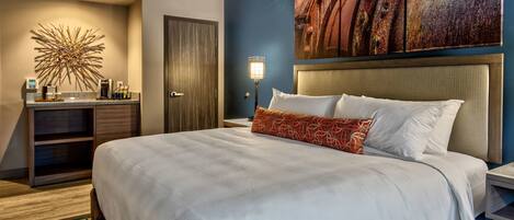 A hotel room with a large bed, a nightstand, a desk with a lamp, a coffee maker, and a decorative wall piece.