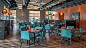 A modern restaurant with wooden flooring, teal chairs, and orange napkins. Tables are set with glasses and cutlery.