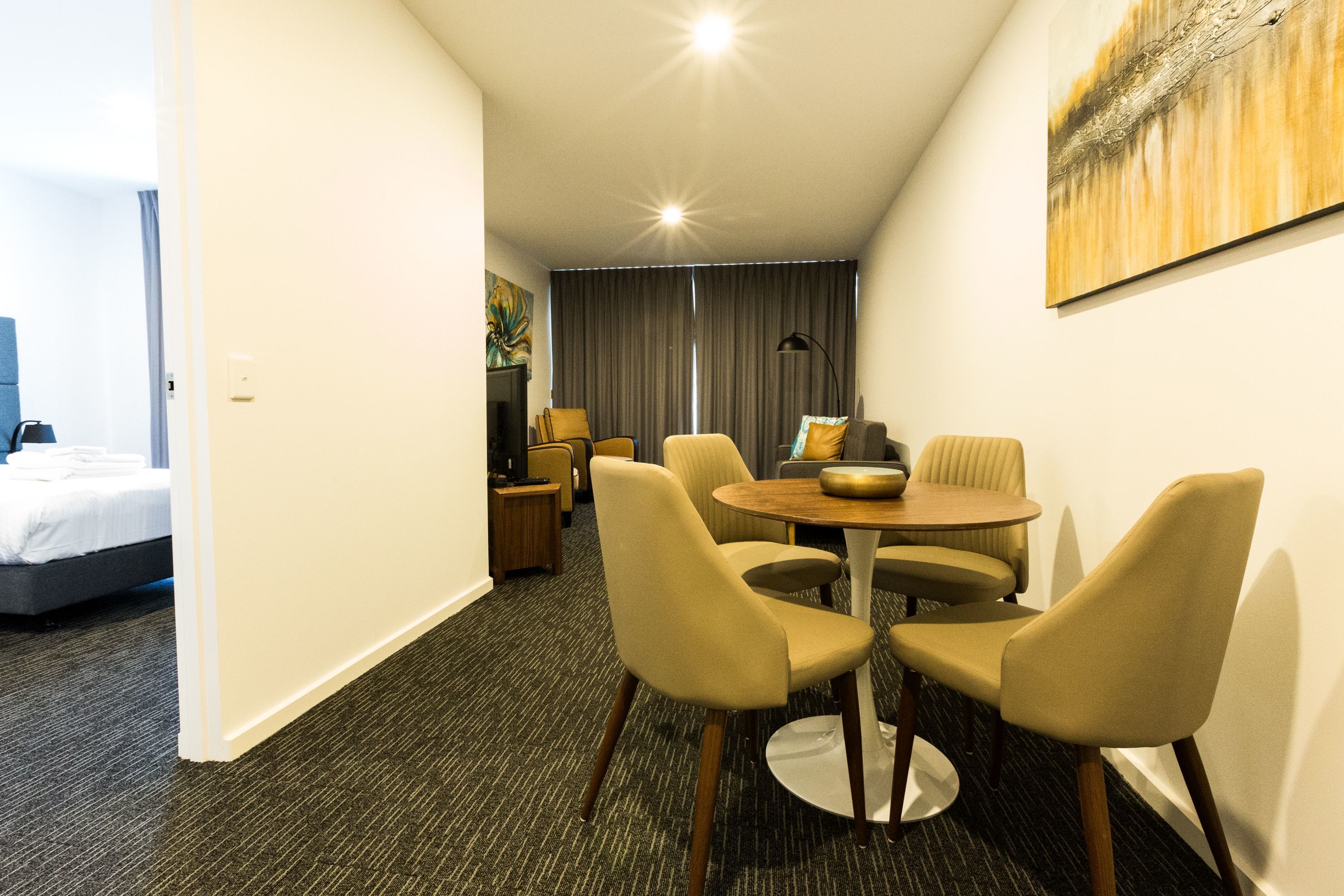 A hotel room with a bed, a dining area with a table and chairs, a lamp, and a painting on the wall.