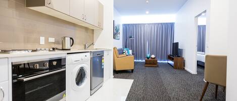A modern kitchen with a washing machine, oven, and a compact dishwasher. There is a living area with a sofa, a coffee table, and a TV. The room has a carpeted floor and a large window with curtains.
