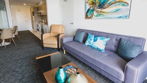 A modern living room with a grey sofa, a beige armchair, and a coffee table with decorative vases. A colorful abstract painting is on the wall, and a kitchen area is visible in the background. - The Palms Apartments (Kent Town)