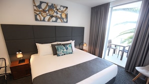 A hotel room with a large bed, a nightstand with a lamp, a chair, and a view of the outdoors. - The Palms Apartments (Kent Town)