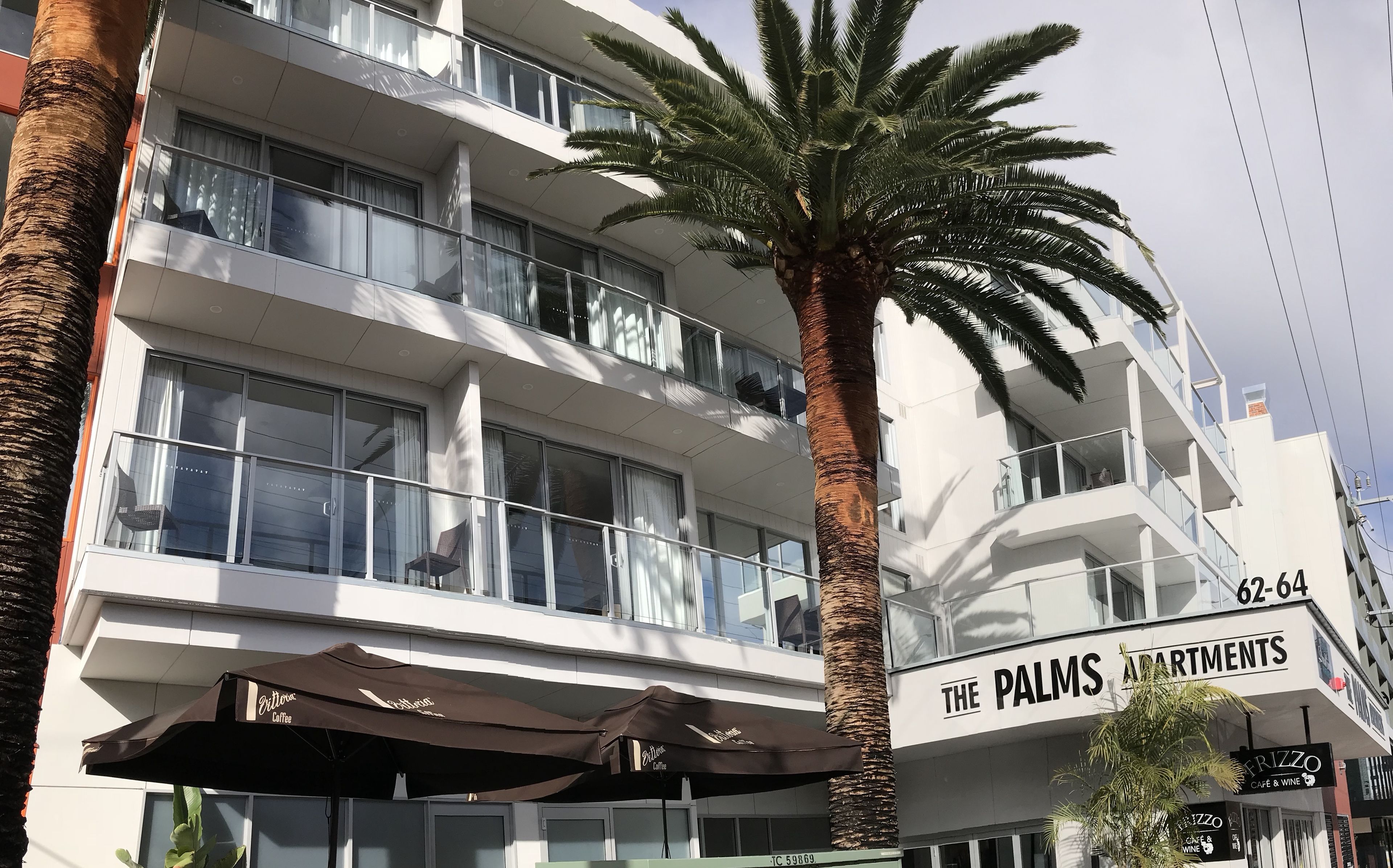 Een meerhoog gebouw met balkons, een bord met 'The Palms Apartments' en een bord met 'Frizzo Cafe & Wine'.