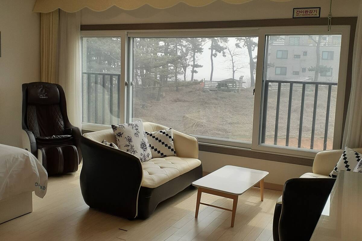 A hotel room with a sofa, armchair, and a small table. There is a large window with a view of trees and buildings outside.
