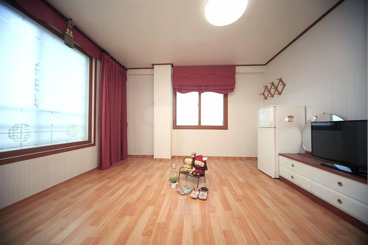 A room with wooden flooring, a television on a wooden stand, a white refrigerator, and a window with red curtains.