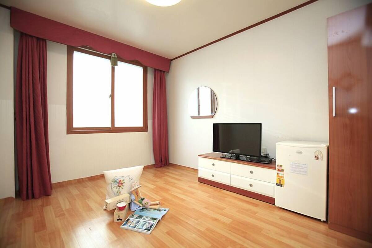 A room with a wooden floor, a television on a white cabinet, a refrigerator, and a window with curtains.