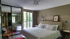 A modern hotel room with a large bed, a sitting area with a chair, a small table, and a large window offering a view of greenery outside. - Aikia City CDMX (Mexico City)
