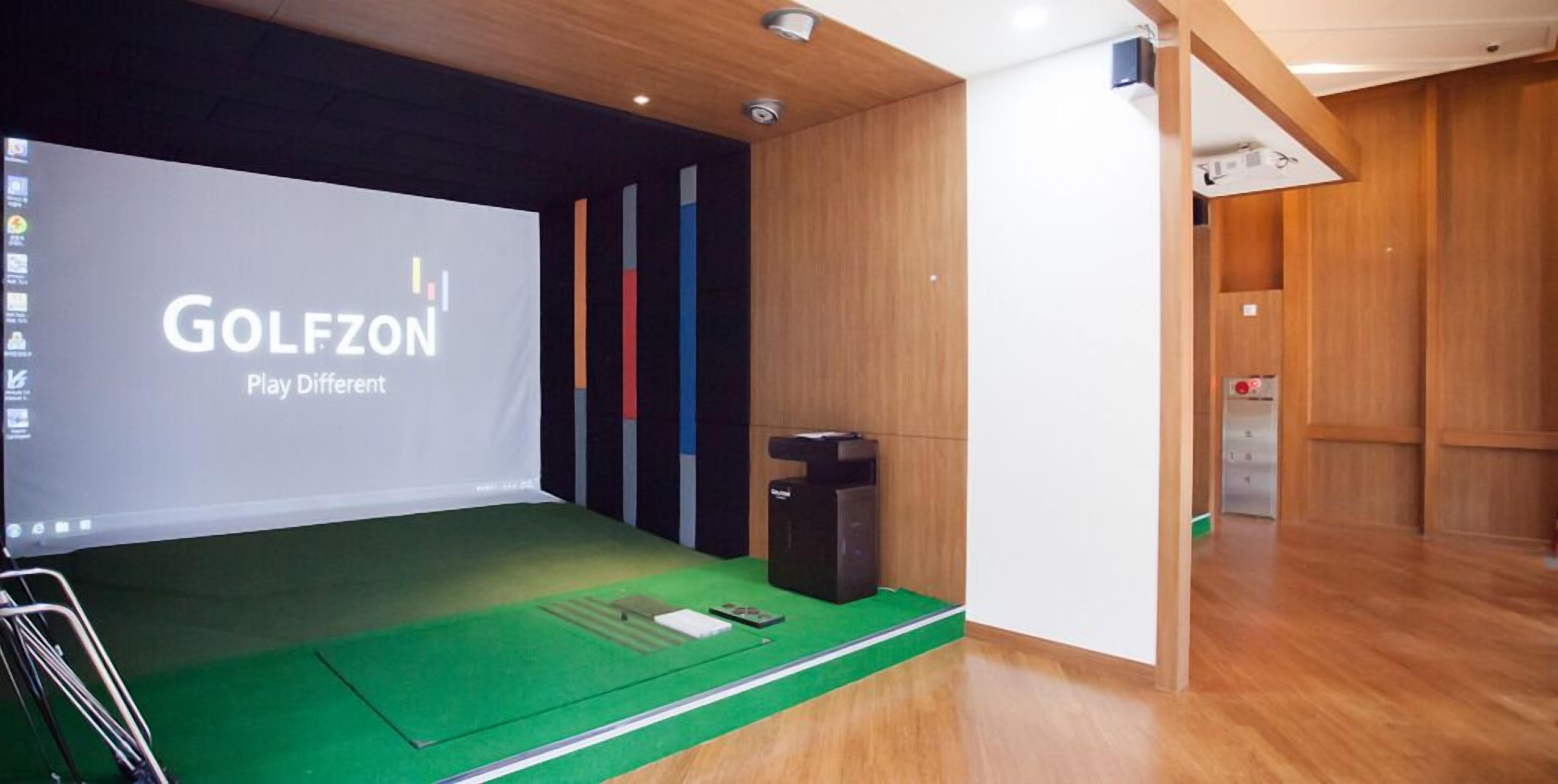 A room with a green carpeted area, a projection screen displaying 'GOLFZONE Play Different', and a wooden panel wall.