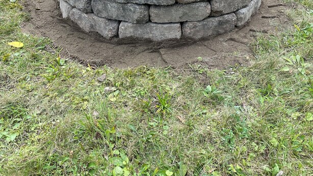 A stone fire pit with a metal rim, surrounded by a grassy area and dense foliage in the background. - A Little Piece of Relaxation (Millport)