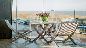 A balcony with a table and two chairs, a vase with flowers, and a view of the beach and parked cars. - Apartment in Egmond with Sea Views (Egmond aan zee)