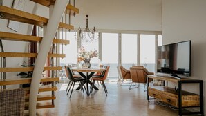 A spacious living area with a dining table, chairs, a TV, and a view of the sea. - Apartment in Egmond with Sea Views (Egmond aan zee)