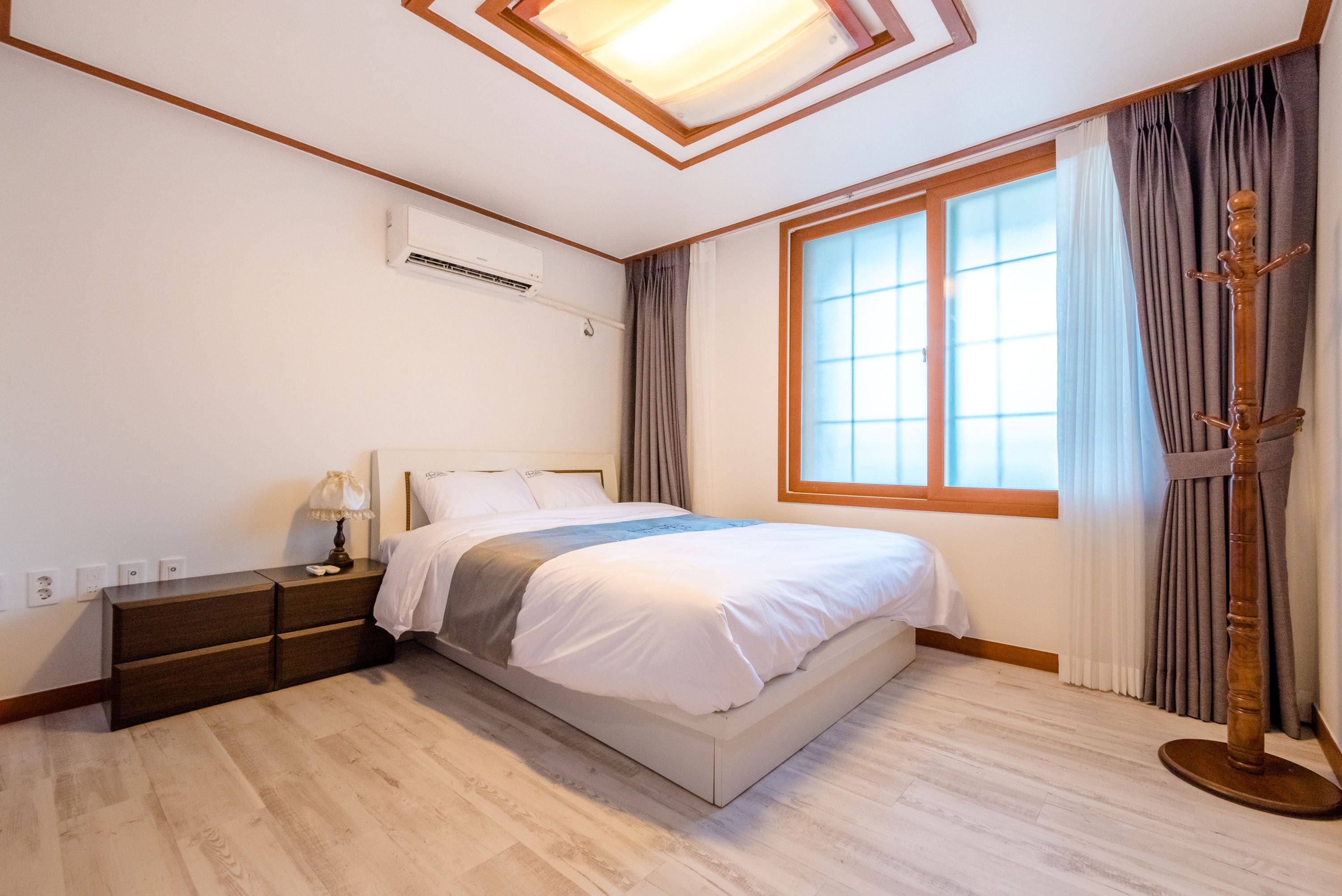 A bedroom with a bed, bedside tables, an air conditioning unit, a window with curtains, and a wooden floor.