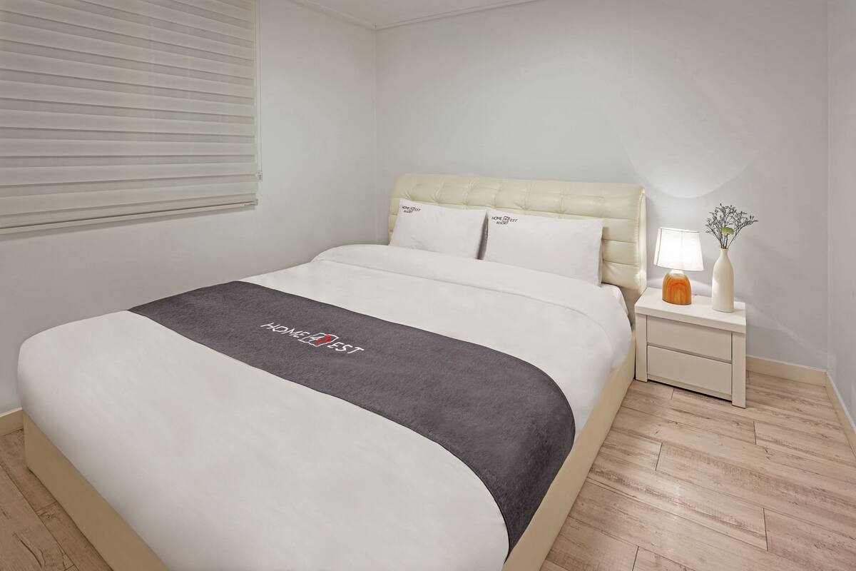 A neatly made bed with a grey and white bedspread, a white headboard, and a bedside table with a lamp and a vase.