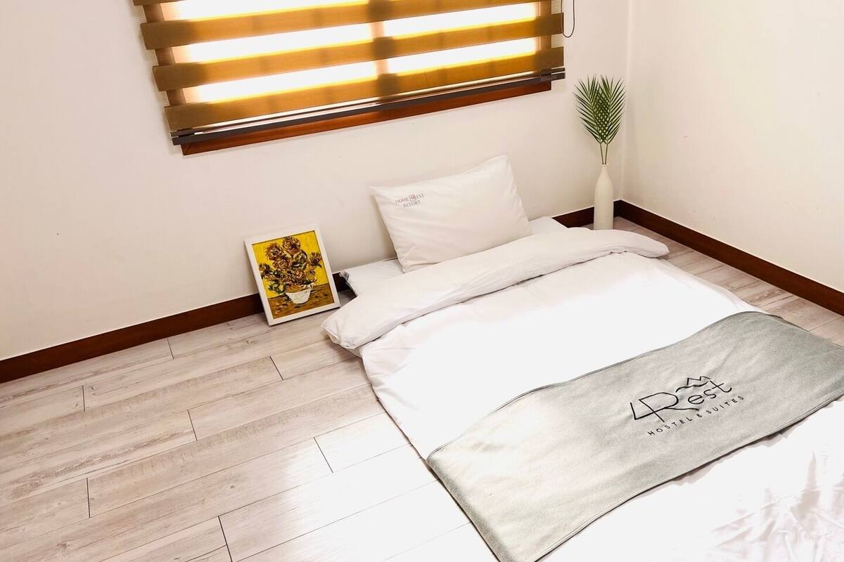 A bedroom with a bed, a bedside table with a plant, a framed picture, and a window with blinds.