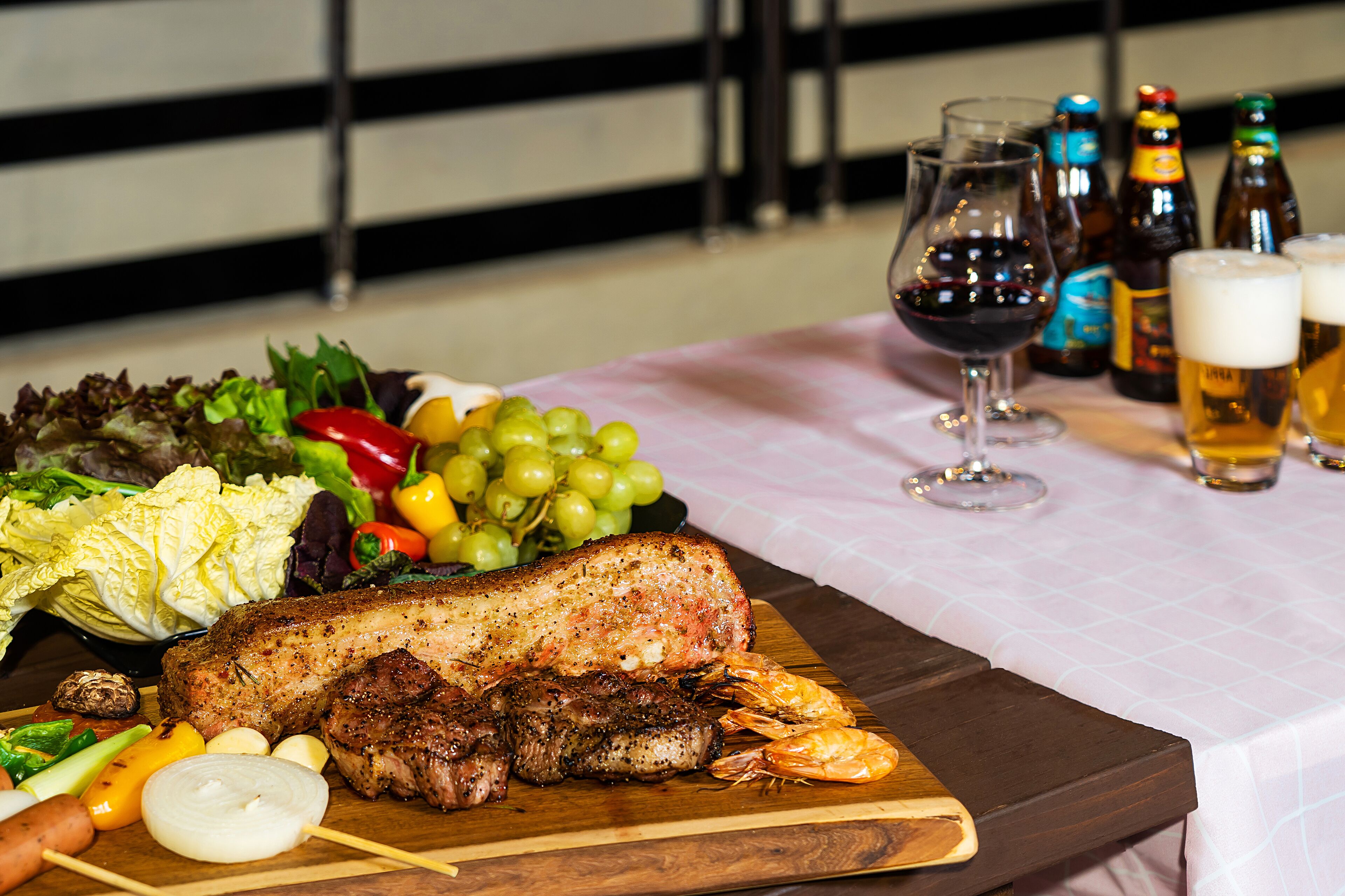 A table with a variety of food including a large piece of meat, shrimp, and vegetables, along with several bottles and glasses of beer.