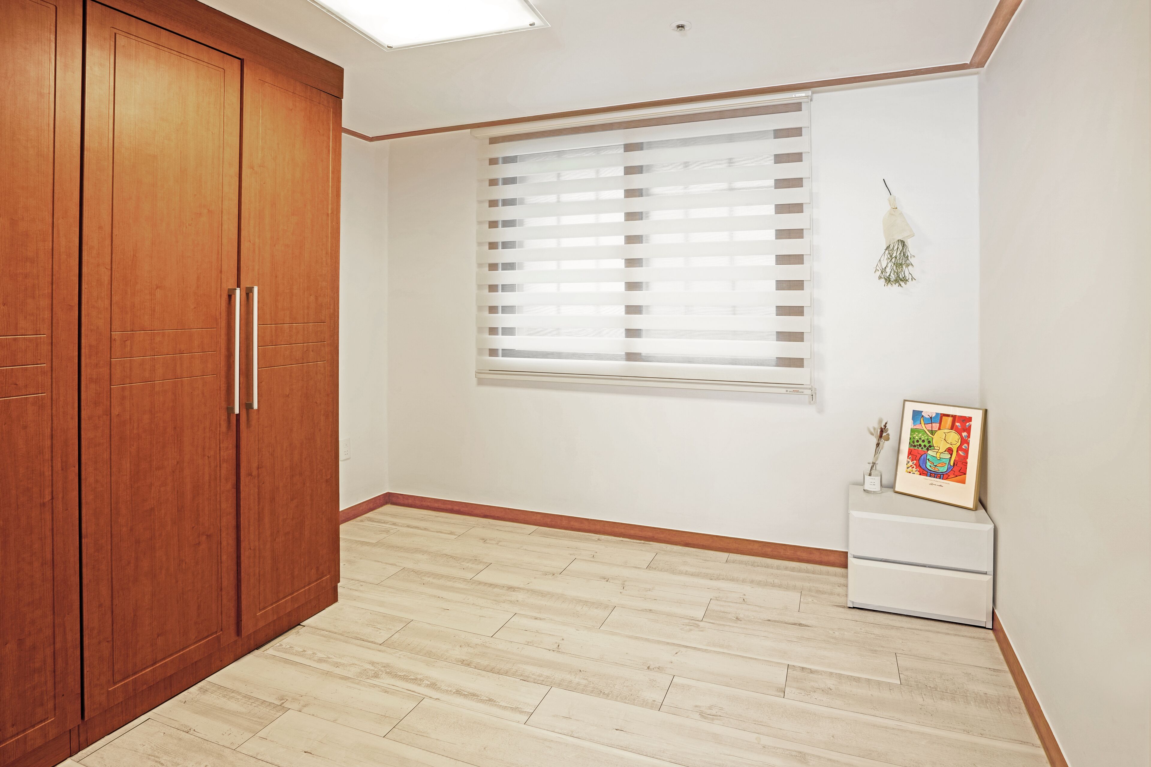 A room with a wooden wardrobe, a window with blinds, a white bedside table with a framed picture, and a light-colored wooden floor.