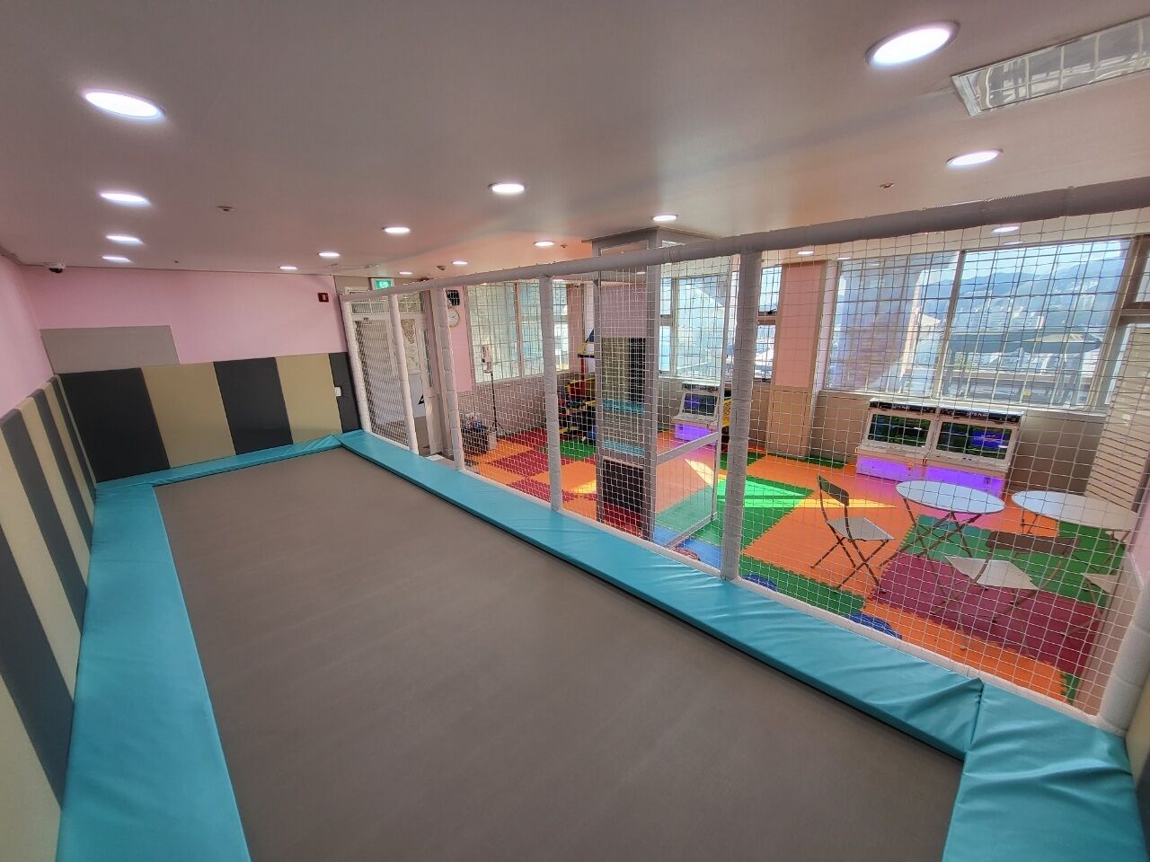 Un gimnasio con zona de suelo acolchado, equipamiento de ejercicio y una área de juegos colorida visible a través de una red de seguridad.