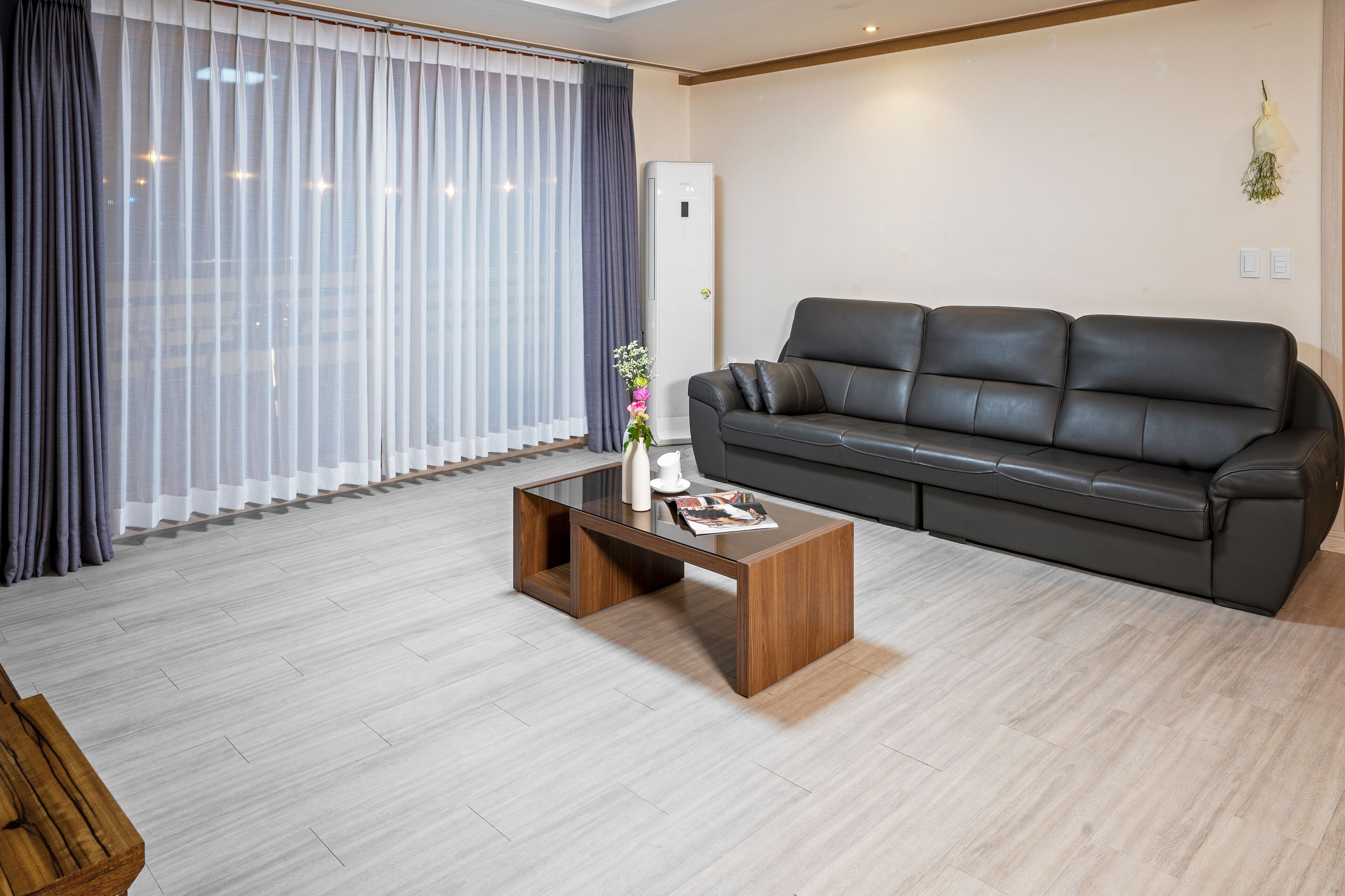 A modern living room with a dark leather sofa, a wooden coffee table, and a large window with sheer curtains.