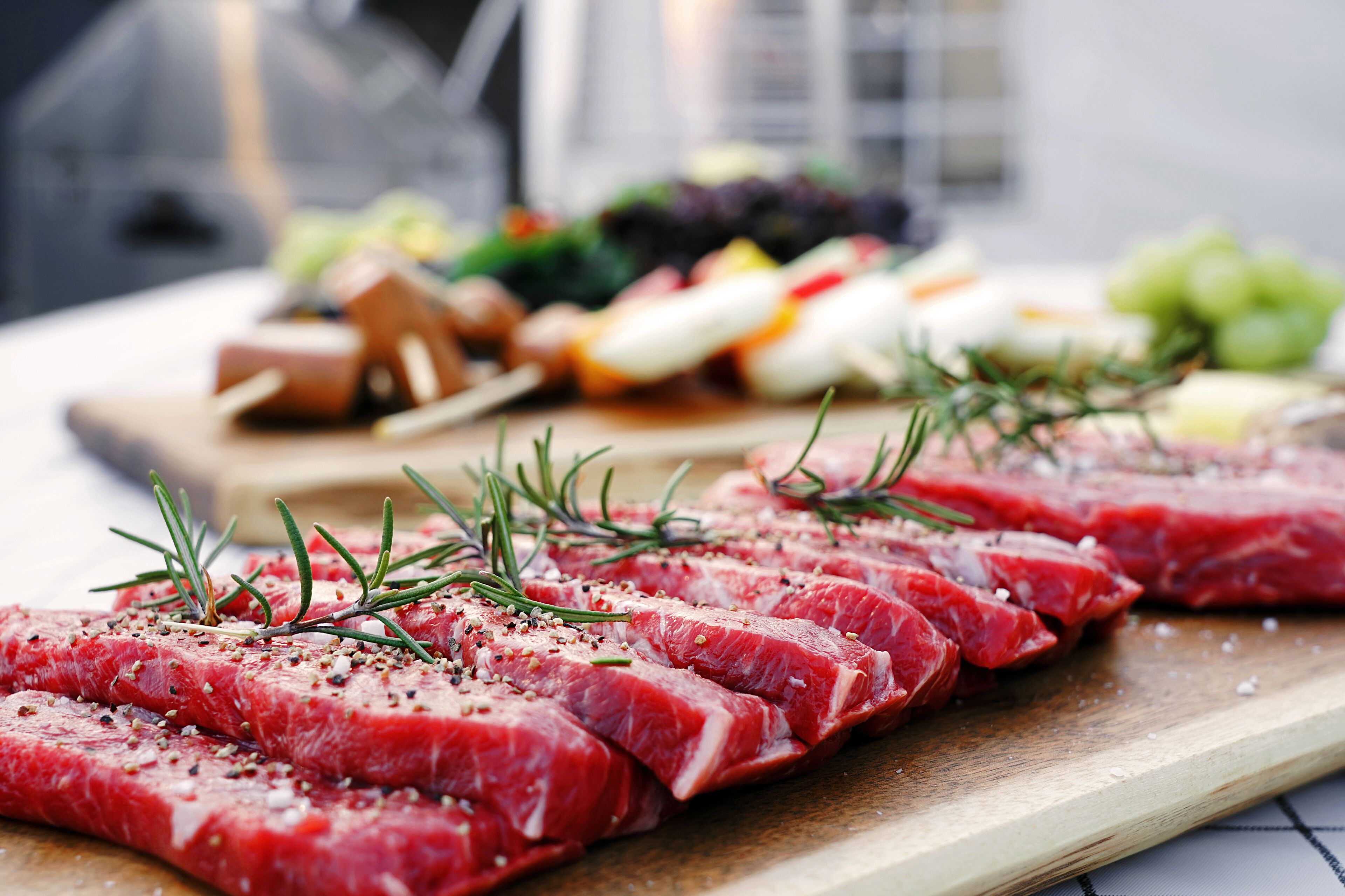 Raw steak slices on a wooden board with rosemary sprigs, accompanied by various cheeses and fruits in the background.