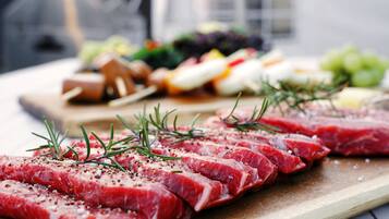 Raw steak slices on a wooden board with rosemary sprigs, accompanied by various cheeses and fruits in the background.