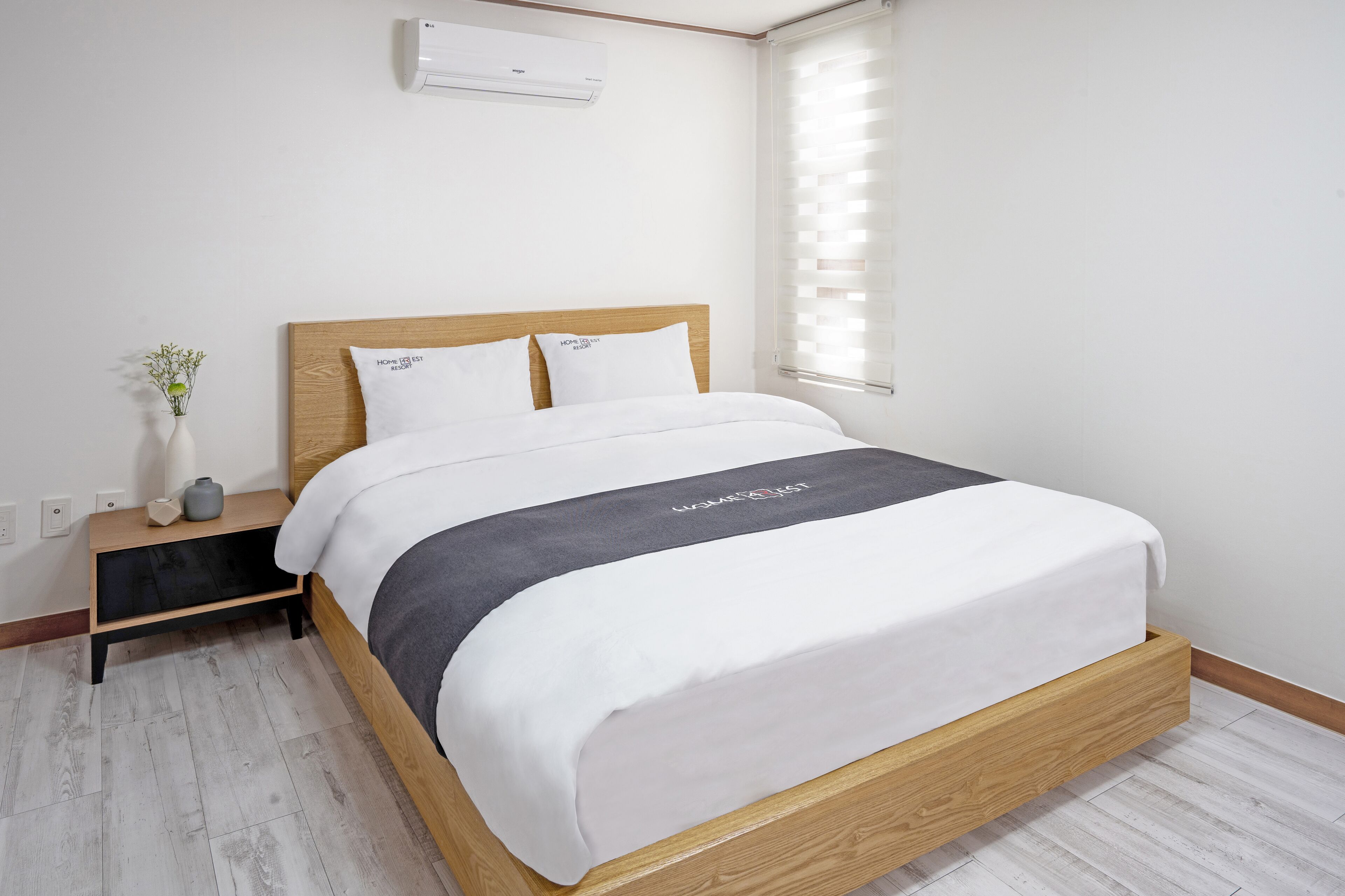 A neatly made bed with a wooden headboard, a bedside table with a vase and a candle, and a window with blinds.