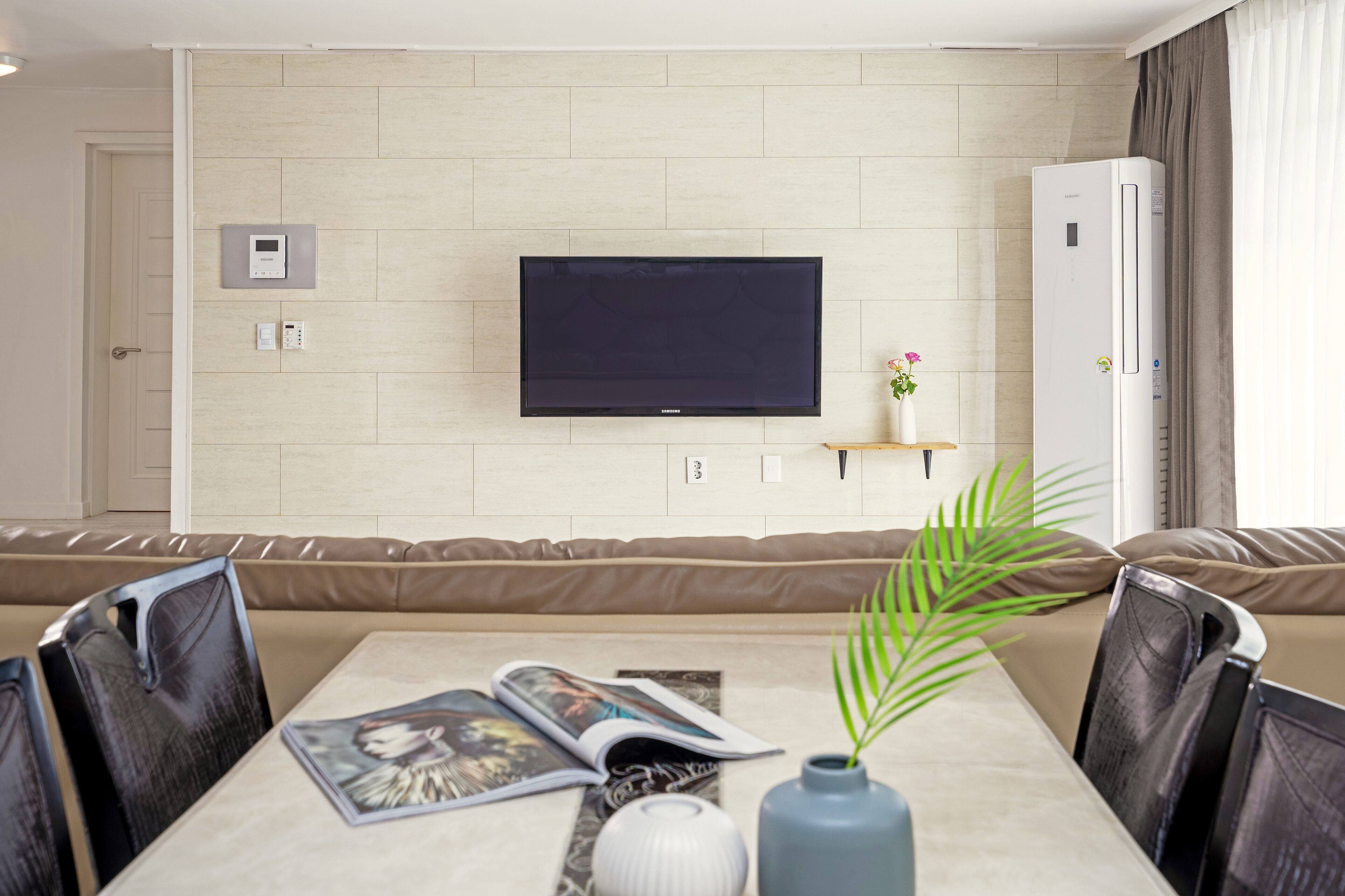 Ein modernes Wohnzimmer mit Flachbildfernseher an der Wand, einer Couch, einem Esstisch mit Stühlen und einer kleinen Pflanze auf dem Tisch.