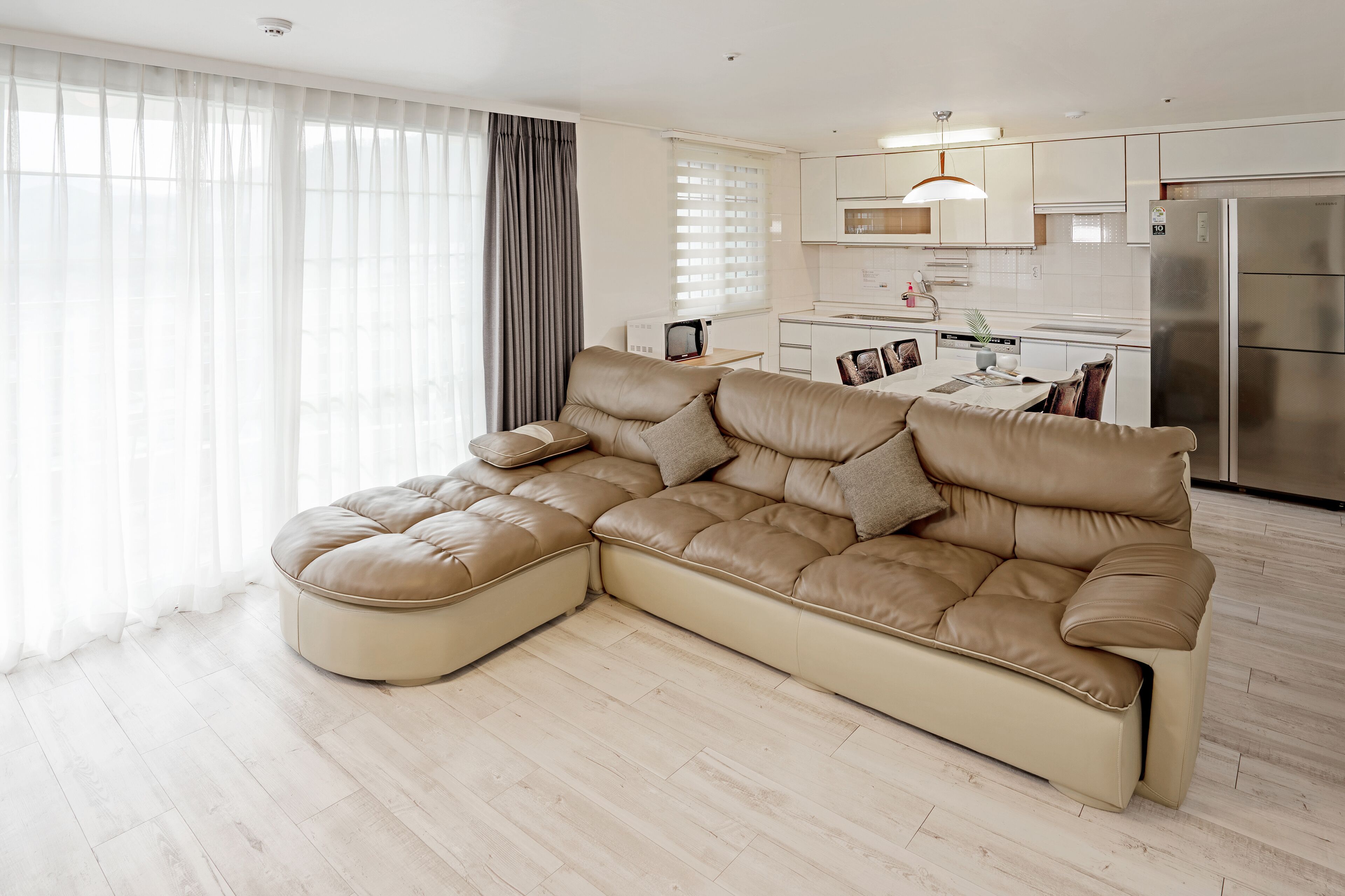 A modern living room with a large L-shaped sectional sofa, a dining table with chairs, and a kitchen area with a refrigerator and microwave.