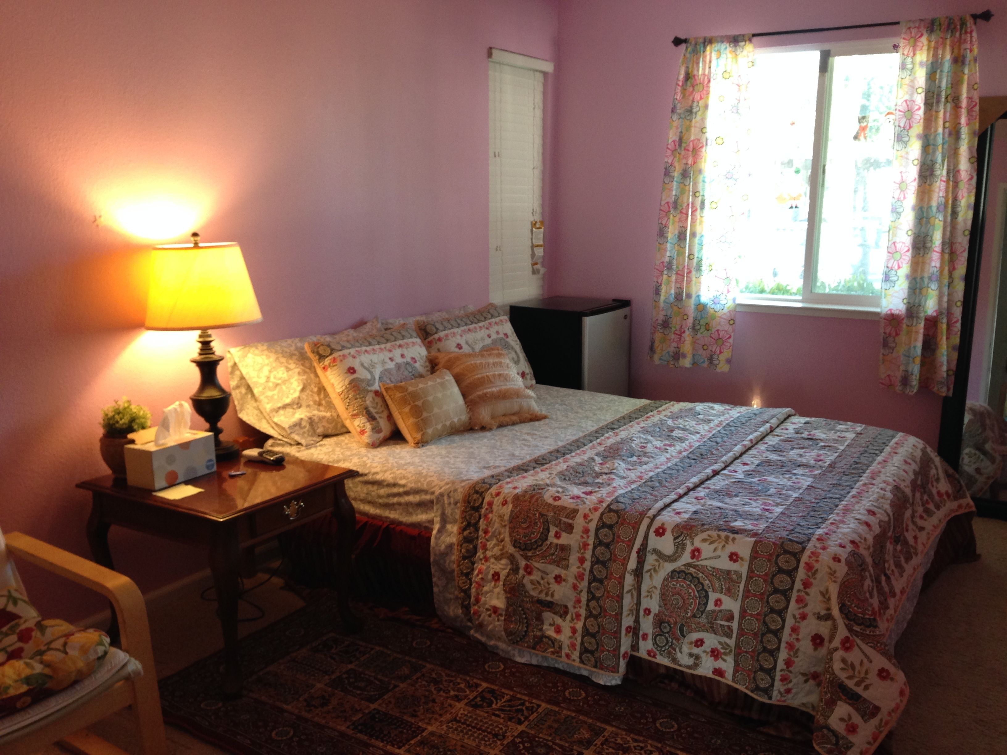 Un dormitorio con cama, mesita de noche, lámpara, silla, ventana con cortinas y puerta.
