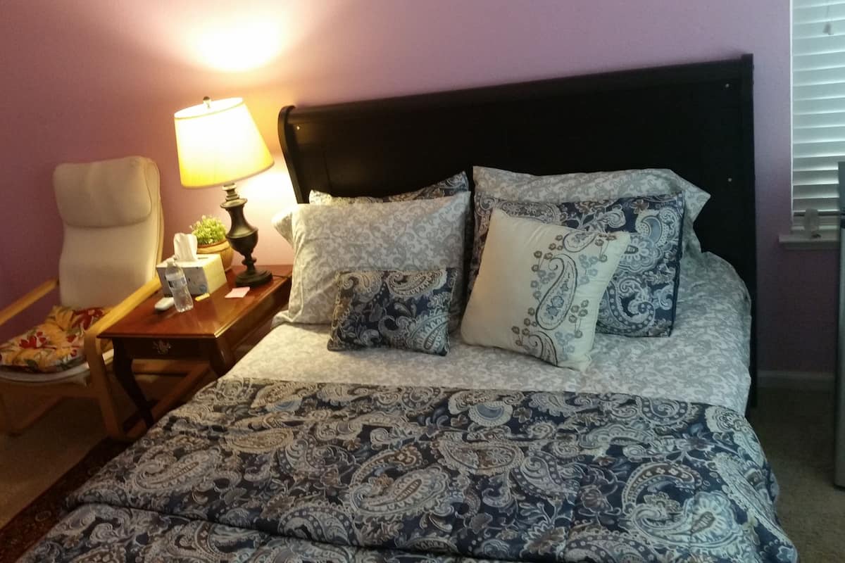 A neatly made bed with patterned bedding, a bedside table with a lamp, a chair, and a small table with a tissue box and water bottle.